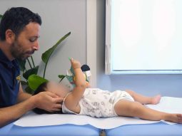 un uomo con una camicia blu navy esamina delicatamente un neonato disteso su un lettino da visita in una stanza ben illuminata con delle piante sullo sfondo.